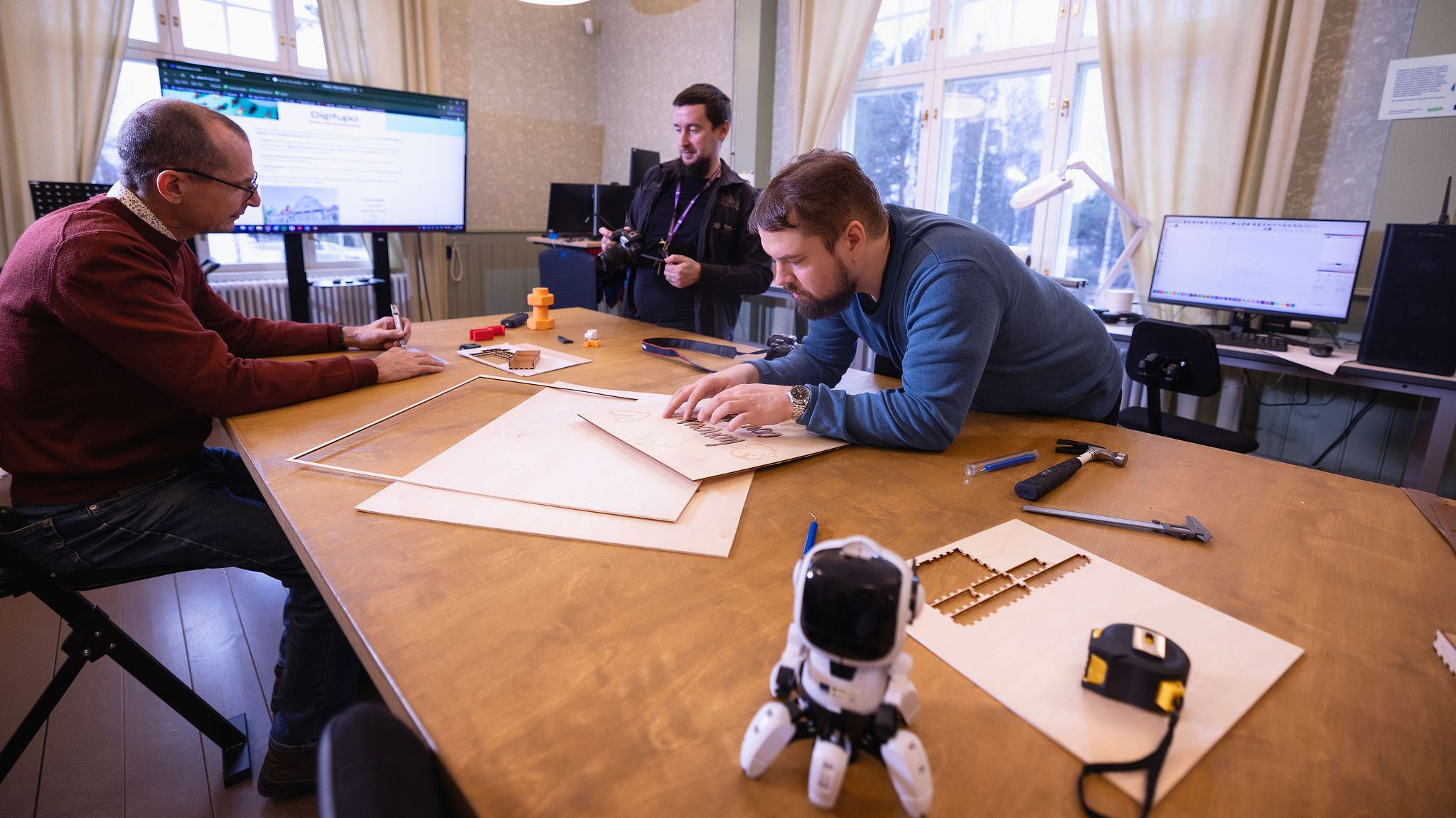 Kolme henkilöä Digituvan pajalla työpöydän ääressä. Pöydällä muun muassa laservaneria, 3D-tulosteita sekä robotti.