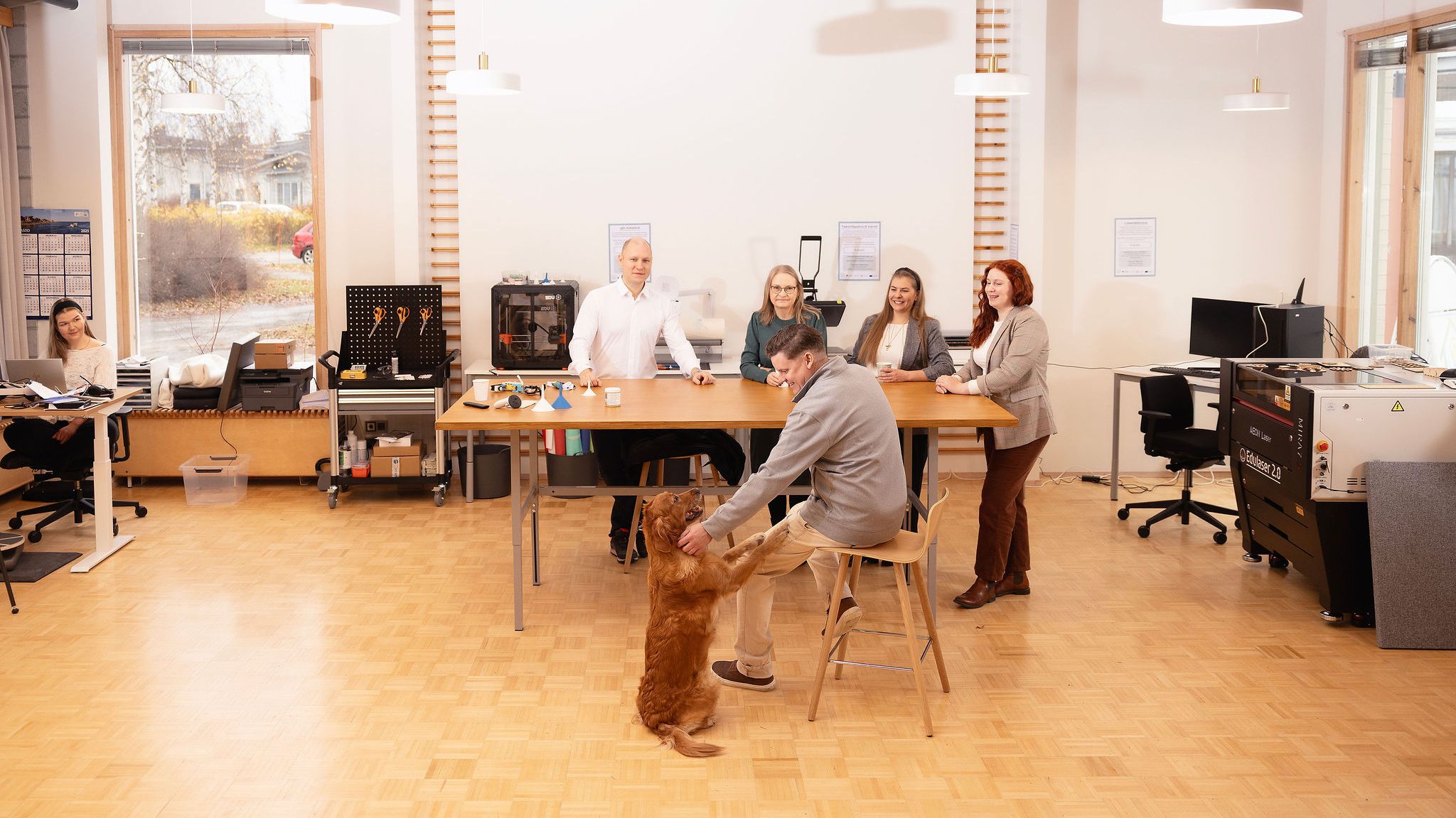 Henkilöt Juuan Digituvan pajalla. Kuvassa näkyy Digituvan laitteistoa, kuten laserleikkuri ja 3D-tulostin. Lisäksi kuvassa kultainen noutaja.