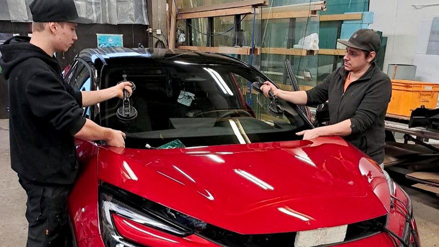 Yrittäjä Ville Pulkkinen ja Jarmo Lohtander asentamassa tuulilasia punaiseen autoon.